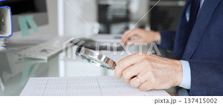 Hand holding magnifying glass, inspecting financial document, performing audit and business analysis. Audit and taxes in business Hand holding magnifying glass, inspecting financial document, performing audit and business analysis. Audit and taxes in business 137415002