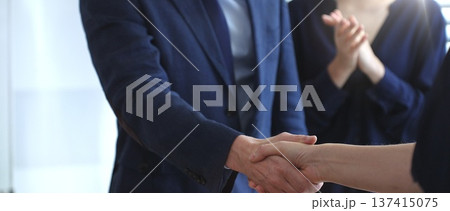 Business people shaking hands during a successful corporate meeting, finalizing an important deal while a colleague claps in approval, symbolizing strong teamwork Business people shaking hands during a successful corporate meeting, finalizing an important deal while a colleague claps in approval, symbolizing strong teamwork 137415075