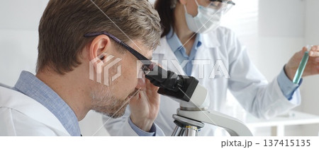 A male researcher examines a sample under a microscope, a female lab technician in a protective mask stands nearby, both in white lab coats during the study. Medicine and Science A male researcher examines a sample under a microscope, a female lab technician in a protective mask stands nearby, both in white lab coats during the study. Medicine and Science 137415135