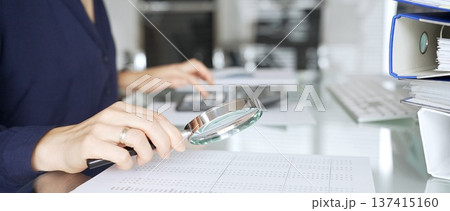 Person auditing financial document using magnifying glass on desk with binders, researching business data. Audit and taxes in business Person auditing financial document using magnifying glass on desk with binders, researching business data. Audit and taxes in business 137415160