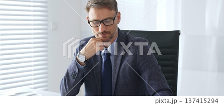 Adult businessman wearing blue suit and tail is concentrating and thinking while working at the desk in a bright, modern office, demonstrating professionalism and efficiency. Business people concept Adult businessman wearing blue suit and tail is concentrating and thinking while working at the desk in a bright, modern office, demonstrating professionalism and efficiency. Business people concept 137415294
