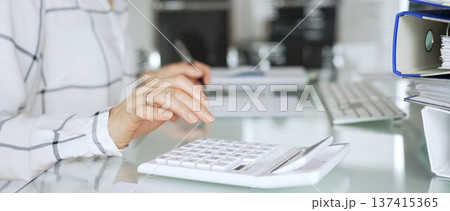 Woman calculating finances on a white calculator, managing budget and paperwork at office desk, preparing taxes. Audit and taxes in business Woman calculating finances on a white calculator, managing budget and paperwork at office desk, preparing taxes. Audit and taxes in business 137415365