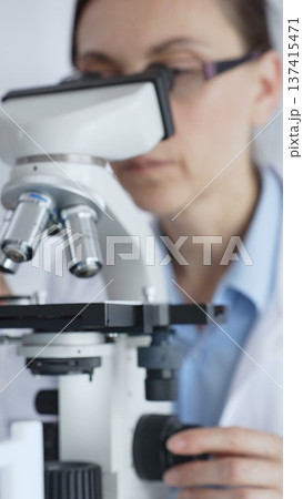 Female scientific researcher wearing protective equipment checking microscope, analyzing blue test tubes in modern laboratory research setting. Medicine and science concept Female scientific researcher wearing protective equipment checking microscope, analyzing blue test tubes in modern laboratory research setting. Medicine and science concept 137415471