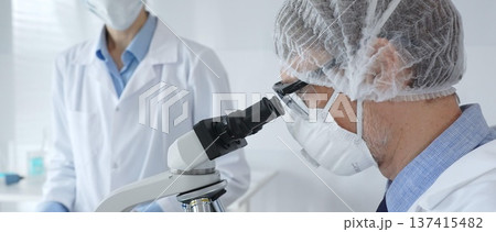 A male scientist wearing a protective mask, glasses and hairnet works with a microscope while a female colleague helps him by holding a tablet and taking notes in a sterile laboratory. Concept of 137415482