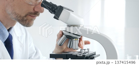 Adult male scientific researcher analyzing sample through microscope, wearing protective gear in high tech laboratory environment. Healthcare, medicine and science concept 137415496