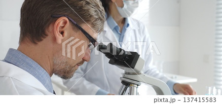 A researcher wearing glasses examines a sample under a microscope, a lab technician wearing a protective mask stands nearby, both in white lab coats during the study. Medicine and Science A researcher wearing glasses examines a sample under a microscope, a lab technician wearing a protective mask stands nearby, both in white lab coats during the study. Medicine and Science 137415583