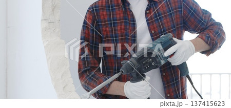 Male construction worker wearing red checkered shirt and protective white gloves is demolishing white wall with rotary hammer drill and generating dust during renovation project, closeup view Male construction worker wearing red checkered shirt and protective white gloves is demolishing white wall with rotary hammer drill and generating dust during renovation project, closeup view 137415623