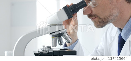 Adult male scientific researcher analyzing sample through microscope, wearing protective gear in high tech laboratory environment. Healthcare, medicine and science concept 137415664