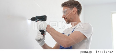 Man professional construction worker in a blue jumpsuit, safety goggles, and work gloves is screwing a screw into the wall using a cordless screwdriver, home renovation Man professional construction worker in a blue jumpsuit, safety goggles, and work gloves is screwing a screw into the wall using a cordless screwdriver, home renovation 137415732