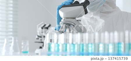 Male doctor in protective suit and gloves using microscope while analyzing samples while conducting research in sterile laboratory. Close-up of test tubes with blue liquid 137415739