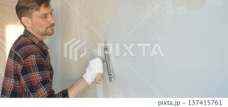 Male construction worker wearing gloves is carefully applying plaster to a wall using a hand tool, contributing to a home renovation or construction project 137415761