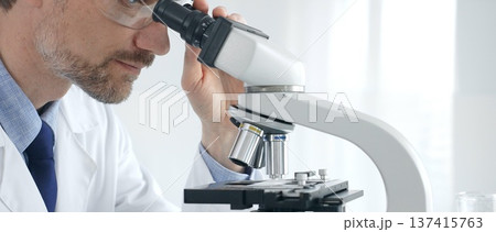 Adult male scientific researcher analyzing sample through microscope, wearing protective gear in high tech laboratory environment. Healthcare, medicine and science concept Adult male scientific researcher analyzing sample through microscope, wearing protective gear in high tech laboratory environment. Healthcare, medicine and science concept 137415763