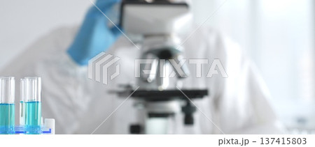 Male medical scientist in protective suit using microscope while analyzing samples, conducting research in laboratory. Close-up of test tubes with blue liquid 137415803