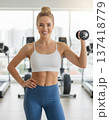 Athletic blond smiling young woman in a white sports top and blue leggings with a small dumbbell in hand in gym background. 137418779