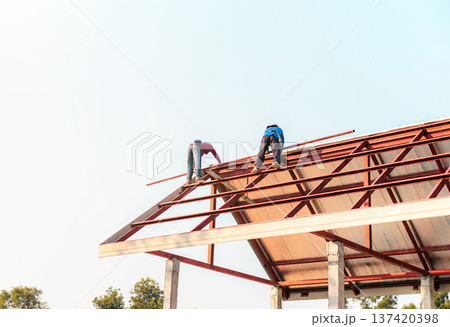 Roofer working in special protective work wear gloves, using air or pneumatic nail gun installing asphalt shingle on top of the new roof, Concept of residential building under construction. Roofer working in special protective work wear gloves, using air or pneumatic nail gun installing asphalt shingle on top of the new roof, Concept of residential building under construction. 137420398