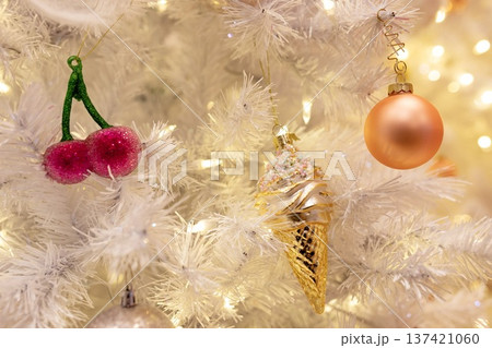 Cute pastel Christmas ornaments, including a pink cherry, golden ice cream cone, and peach bauble, hanging on a white tree with warm glowing lights. A whimsical and festive holiday close-up. Cute pastel Christmas ornaments, including a pink cherry, golden ice cream cone, and peach bauble, hanging on a white tree with warm glowing lights. A whimsical and festive holiday close-up. 137421060