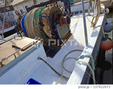 Bright fishing boat with nets on mechanical reel rests at a busy harbor. The weathered deck shows signs of use from daily fishing routines and activities. Bright fishing boat with nets on mechanical reel rests at a busy harbor. The weathered deck shows signs of use from daily fishing routines and activities. 137422673