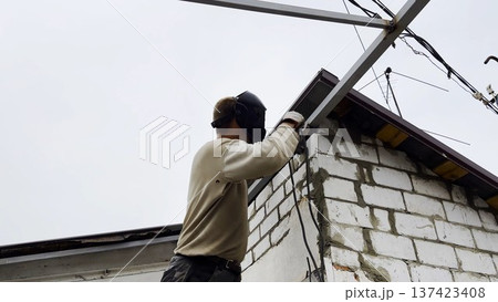Manual laborer in protective mask welds iron detail at construction. Professional young repairman with welding machine working at the site. Male welder operating with metal on house. Close up Manual laborer in protective mask welds iron detail at construction. Professional young repairman with welding machine working at the site. Male welder operating with metal on house. Close up 137423408
