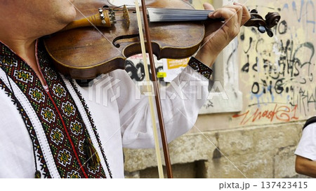 Adult man wearing a traditional embroidered vyshyvanka plays a vintage violin on city street. The scene emphasizes cultural identity, heritage and the expressive character of live music. Slow mo Adult man wearing a traditional embroidered vyshyvanka plays a vintage violin on city street. The scene emphasizes cultural identity, heritage and the expressive character of live music. Slow mo 137423415