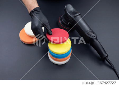 Gloved hand selecting red foam polishing pad near car buffer on dark surface 137427727