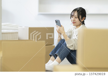 段ボール箱に囲まれて座ってスマートフォンを見る若い女性　引っ越し　イメージ 137429019
