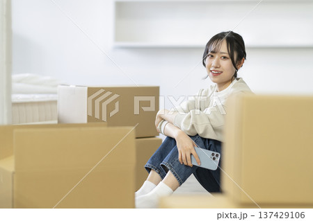 段ボール箱に囲まれて座るスマートフォンを持った若い女性　カメラ目線　引っ越し　イメージ 137429106