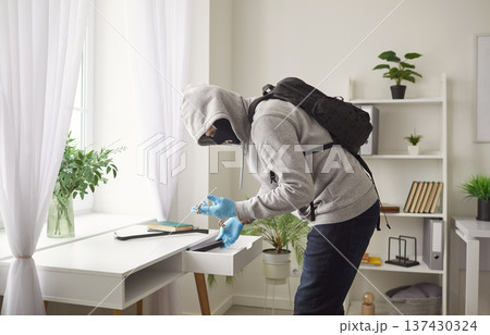Masked burglar in hoodie and gloves searching drawer and holding stolen valuables during robbery. Masked burglar in hoodie and gloves searching drawer and holding stolen valuables during robbery. 137430324