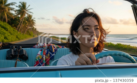 Asian woman driving vintage blue convertible car on tropical coastal highway during golden hour sunset with palm trees and ocean view 137430359