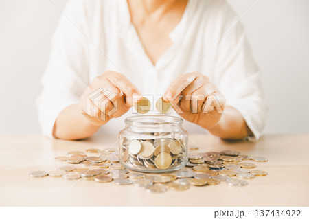 Close-up of hand putting coin into clear glass jar filled with change on bright desk. Minimal home scene for personal savings, budgeting, money management, future goals and security. money management Close-up of hand putting coin into clear glass jar filled with change on bright desk. Minimal home scene for personal savings, budgeting, money management, future goals and security. money management 137434922