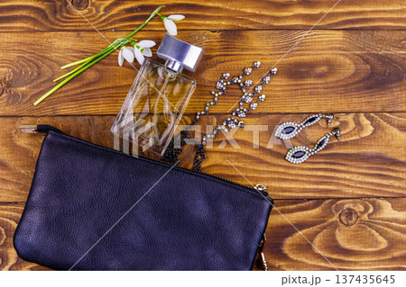 Women accessories on wooden background. Clutch bag, bottle of perfume, necklace, earrings and snowdrops on wood table. Beauty and fashion composition. Top view, flat lay Women accessories on wooden background. Clutch bag, bottle of perfume, necklace, earrings and snowdrops on wood table. Beauty and fashion composition. Top view, flat lay 137435645