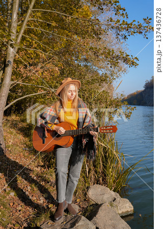 Portrait of smiling caucasian artist plays wooden acoustic guitar in autumnal park. Young songwriter plays string musical instrument hobby outside in nature fall time. Audio music healing Portrait of smiling caucasian artist plays wooden acoustic guitar in autumnal park. Young songwriter plays string musical instrument hobby outside in nature fall time. Audio music healing 137436728
