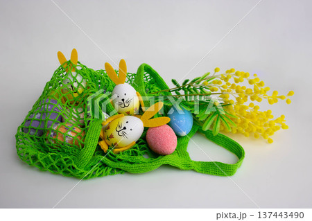 Easter eggs with bunny ears and artificial mimosa branches are lying on the table in a green net. The concept is the Orthodox holiday of Bright Easter 137443490