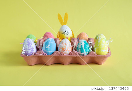 On a yellow background, there is a container with Easter eggs of different colors, including one egg with rabbit ears. 137443496