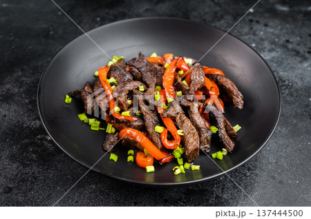 Asian beef stir fry with sweet peppers and scallions in black plate. Pepper steak style beef strips dish on dark background.. Asian beef stir fry with sweet peppers and scallions in black plate. Pepper steak style beef strips dish on dark background.. 137444500