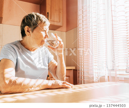 Elderly caucasian woman enjoys a glass of water in sunlit kitchen 137444503
