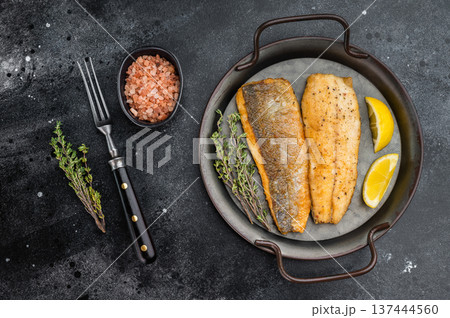Fried sea bass fillets served on ceramic plate with fork and herbs, overhead restaurant style seafood dish ready to eat with dramatic dark backdrop. 137444560