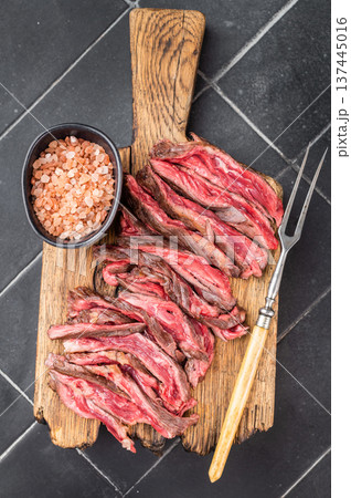 Grilled beef skirt steak slices with coarse salt on wooden cutting board dark textured kitchen background overhead gourmet serving style Grilled beef skirt steak slices with coarse salt on wooden cutting board dark textured kitchen background overhead gourmet serving style 137445016