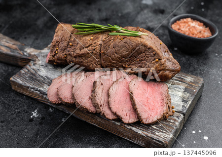 Gourmet roasted beef tenderloin sliced and served with rosemary salt and knife on dark textured background copy space Gourmet roasted beef tenderloin sliced and served with rosemary salt and knife on dark textured background copy space 137445096