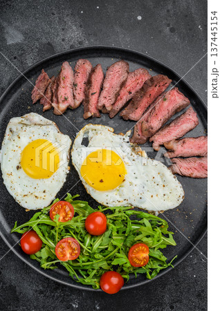 Hearty American steak and eggs with grilled beef, fried eggs and arugula salad, overhead food photo on dark concrete background with copy space Hearty American steak and eggs with grilled beef, fried eggs and arugula salad, overhead food photo on dark concrete background with copy space 137445154