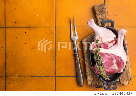 Raw duck legs in a black cast iron skillet, garnished with fresh thyme, resting on a rustic wooden cutting board against an orange tiled background. 137445437