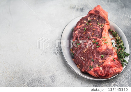Uncooked strip loin new york beef steak with herbs and coarse salt on a silver plate beside vintage carving fork on soft gray textured background. 137445550