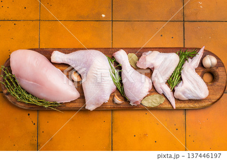 Fresh assortment of chicken breast, wings, drumsticks, and leg quarters on a wooden cutting board with spices, set on an orange kitchen counter. Fresh assortment of chicken breast, wings, drumsticks, and leg quarters on a wooden cutting board with spices, set on an orange kitchen counter. 137446197