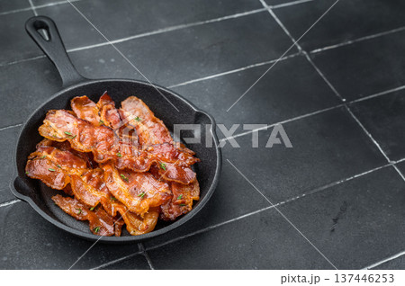Sizzling bacon slices with rich glaze arranged in black skillet against dark tile background delivering bold gourmet look. Sizzling bacon slices with rich glaze arranged in black skillet against dark tile background delivering bold gourmet look. 137446253