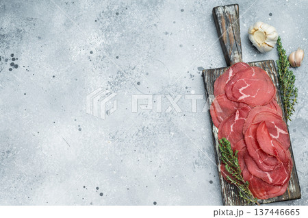 Air dried beef Bresaola charcuterie with marbled red slices, herbs and garlic on a vintage board, top view on a clean stone surface. Air dried beef Bresaola charcuterie with marbled red slices, herbs and garlic on a vintage board, top view on a clean stone surface. 137446665