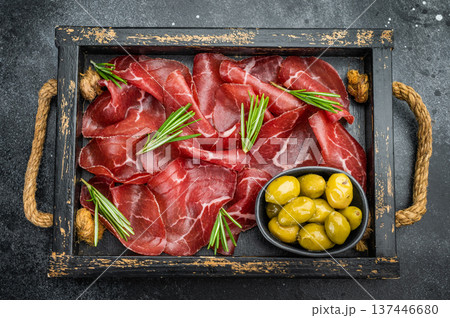 Italian bresaola with rosemary and green olives served in a vintage tray on a dark textured background, shot overhead for gourmet styling. 137446680