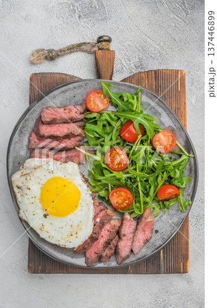 Classic American steak and eggs featuring medium rare beef steak, sunny side egg and fresh greens on wooden board, overhead concrete background Classic American steak and eggs featuring medium rare beef steak, sunny side egg and fresh greens on wooden board, overhead concrete background 137446899