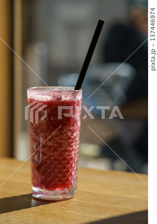 Refreshing red berry smoothie with ice in textured glass and straw on wooden table. Healthy fruit drink served indoors at cozy cafe with soft light Refreshing red berry smoothie with ice in textured glass and straw on wooden table. Healthy fruit drink served indoors at cozy cafe with soft light 137446974