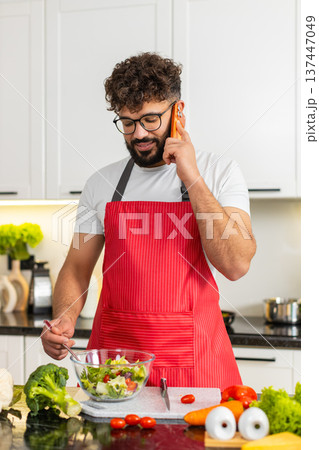 Arabian young adult man mixing salad and talking on phone in kitchen sharing healthy diet fit recipe 137447049