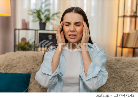 Young woman sitting on sofa holding head suffering from strong headache feeling uncomfortable pain Young woman sitting on sofa holding head suffering from strong headache feeling uncomfortable pain 137447142