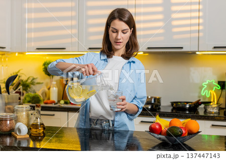 Young woman pouring cold lemon water in home kitchen to quench thirst for diet hydration fit routine Young woman pouring cold lemon water in home kitchen to quench thirst for diet hydration fit routine 137447143